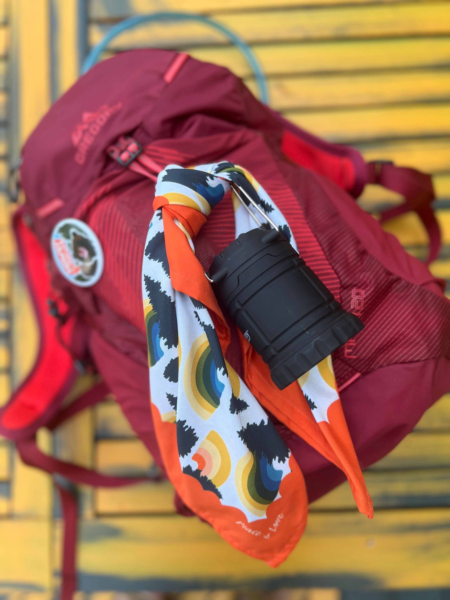 This is a photo showing the Rainbow Pine Bandana tied to a backpack. It's sitting on a yellow picnic table.