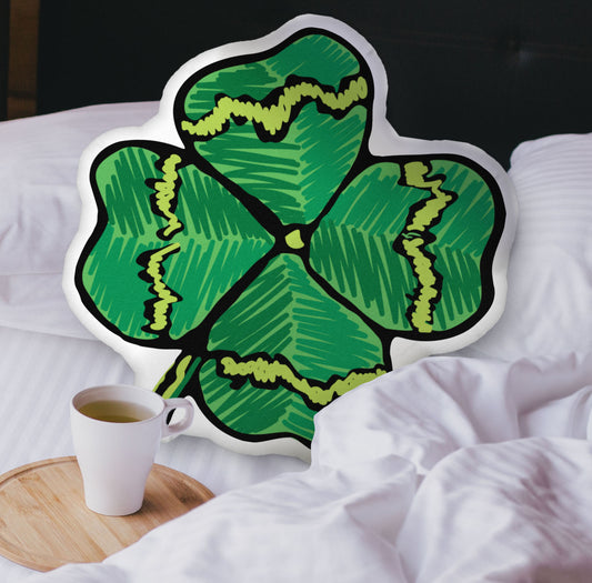 This photograph shows the four-leaf clover-shaped pillow nestled in the white covers of a bed. There is a black headboard and a wooden tray with a coffee mug on the bed.