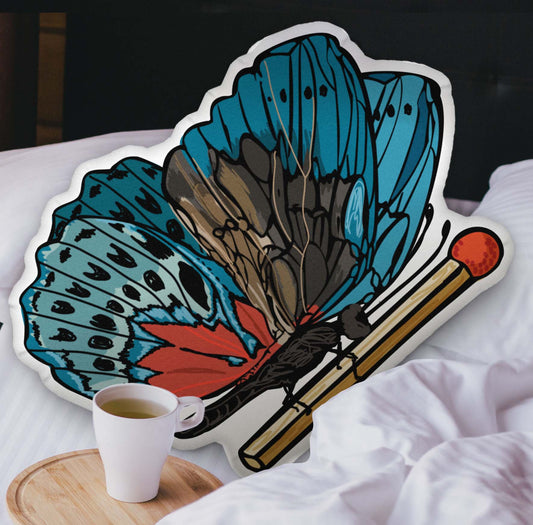 This photograph shows the butterfly-shaped pillow nestled in the white covers of a bed. There is a black headboard and a wooden tray with a coffee mug on the bed.