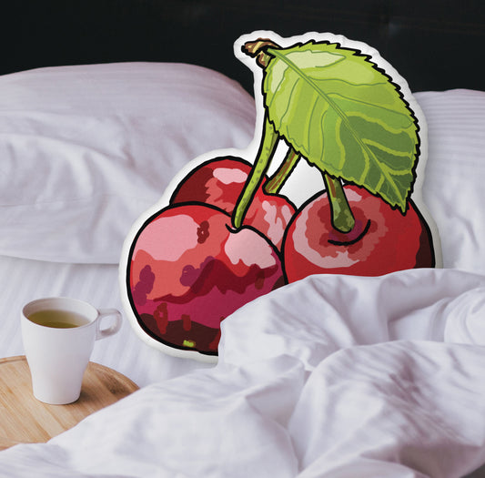 This is a photograph showing the cherry-shaped pillow nestled in the white covers of a bed. There is a black headboard and a wooden tray with a coffee mug on it on the bed.