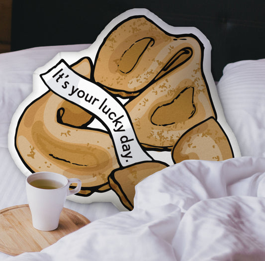 This photograph shows the fortune cookie-shaped pillow nestled in the white covers of a bed. There is a black headboard and a wooden tray with a coffee mug on the bed.