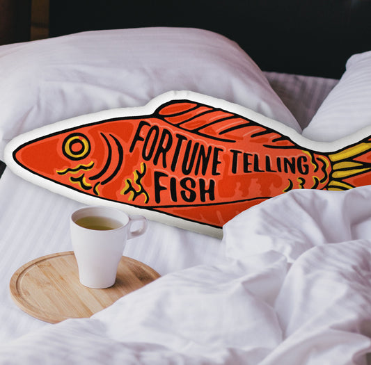 This photograph shows the fish-shaped pillow nestled in the white covers of a bed. There is a black headboard and a wooden tray with a coffee mug on the bed.