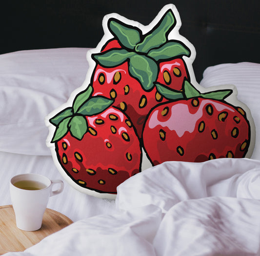 This is a photograph showing the strawberry-shaped pillow nestled in the white covers of a bed. There is a black headboard and a wooden tray with a coffee mug on it on the bed.
