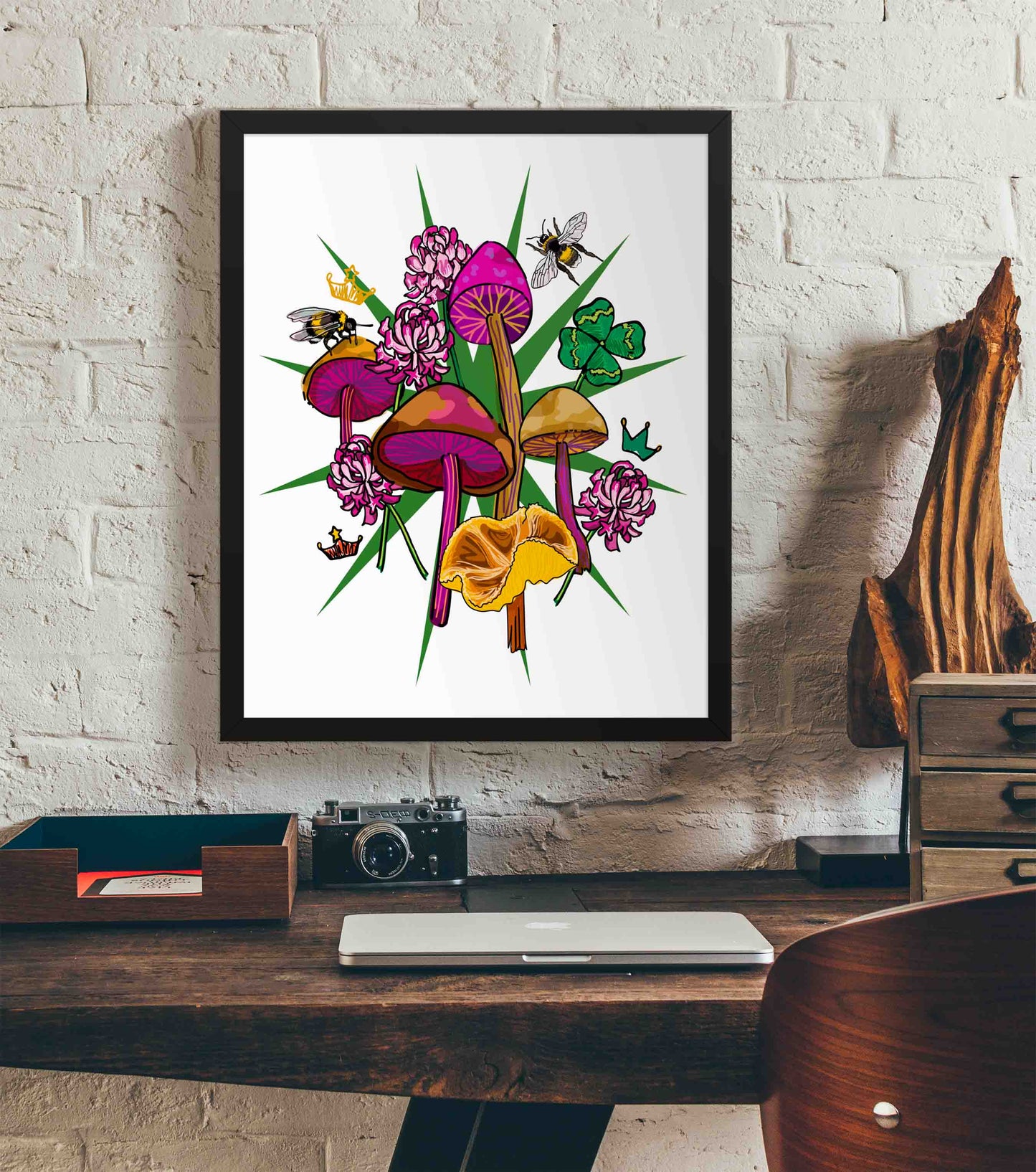 This photo shows the framed Queen's Favor art print hanging on a white brick wall. Below it, there is a wood. The desk has a camera, laptop, and wood sculpture on it.