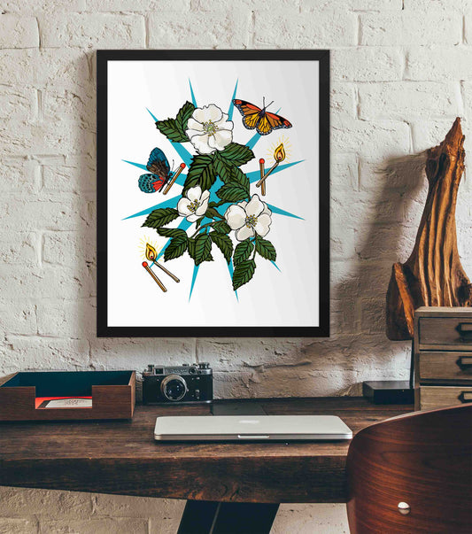 This photo shows the framed Wild Fortune art print hanging on a white brick wall. Below it, there is a wood. The desk has a camera, laptop, and wood sculpture on it.