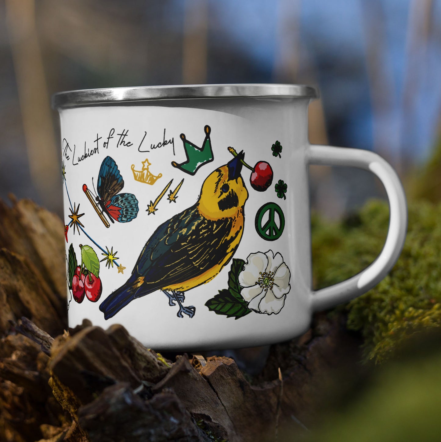 Photo of the Aquarius Stars Camp Mug sitting on a tree stump in the woods. The base color of the enamel cup is white and the artwork is hand drawn illustrations of the Aquarius constellation, known as the "Luckiest of the Lucky" stars. Alongside the stars are other symbols of good luck including a golden finch wearing a crown and eating a cherry, wild roses, peace signs, falling stars, and a butterfly carrying a match. The words "The Luckiest of the Lucky" are woven into the design.