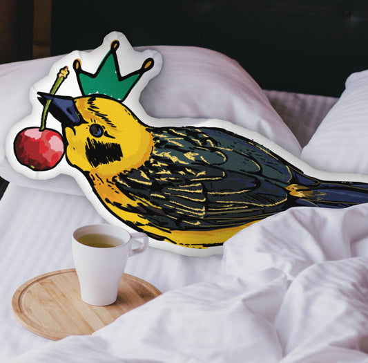 This photograph shows the bird-shaped pillow nestled in the white covers of a bed. There is a black headboard and a wooden tray with a coffee mug on the bed.