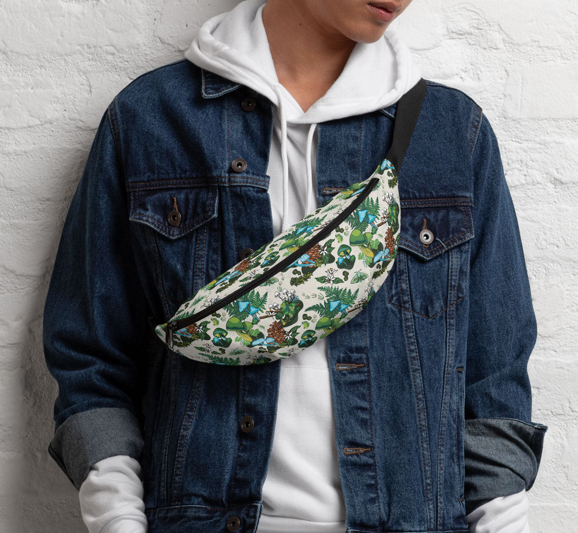 Woman standing against a white brick wall. She is wearing a white sweat shirt under a denim jacket. She is wearing the hip pack like a sling bag over her shoulder.