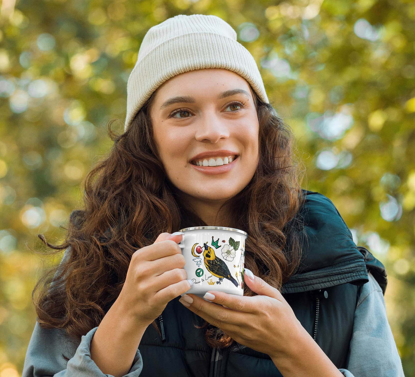 Photo of a female hiker holding the Aquarius Stars Enamel Coffee Cup while standing in the woods. She's wearing a puffer vest over a button-up shirt and an ivory knit cap.