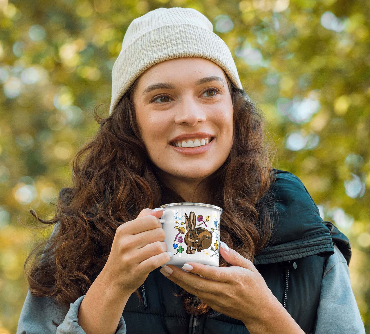 A female hiker is holding the Orion camp mug in the woods. She is wearing an ivory knit cap and a puffer vest over a denim shirt.