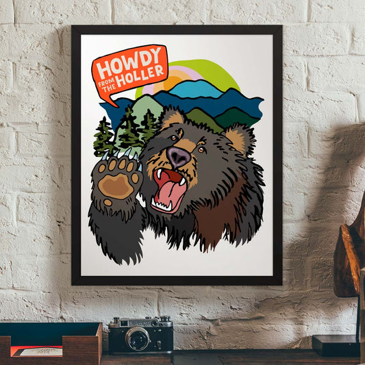 Framed artwork of an illustrated bear in the Appalachian mountains and 'Howdy from the Holler' text. The art print is in a black wood frame and hung on a white brick wall above a desk with a vintage camera.