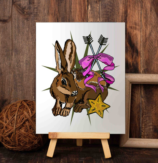 Lucky Rabbit's Path art poster on a wooden easel set against a wood-paneled wall. The art has a multi-pointed star in the background. On top of that, there is feisty bunny, who represents agility, luck, and intuition. The crossed arrows tied with a ribbon symbolize loyalty and friendship. In the lower corner, there is a golden lucky star.