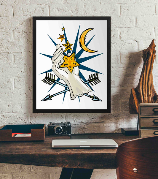 This photo shows the framed Lucky Star Giver art print hanging on a white brick wall. Below it, there is a wood. The desk has a camera, laptop, and wood sculpture on it.