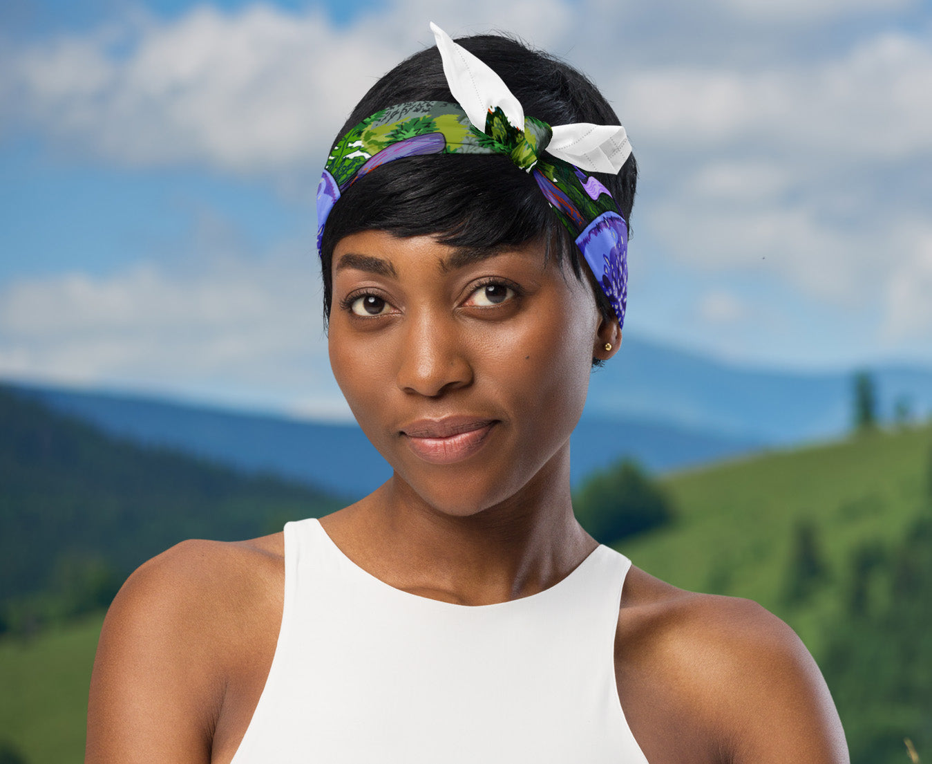 This is an image of a woman wearing the purple mushroom scarf tied around her head as a headband standing in front of a mountain range.