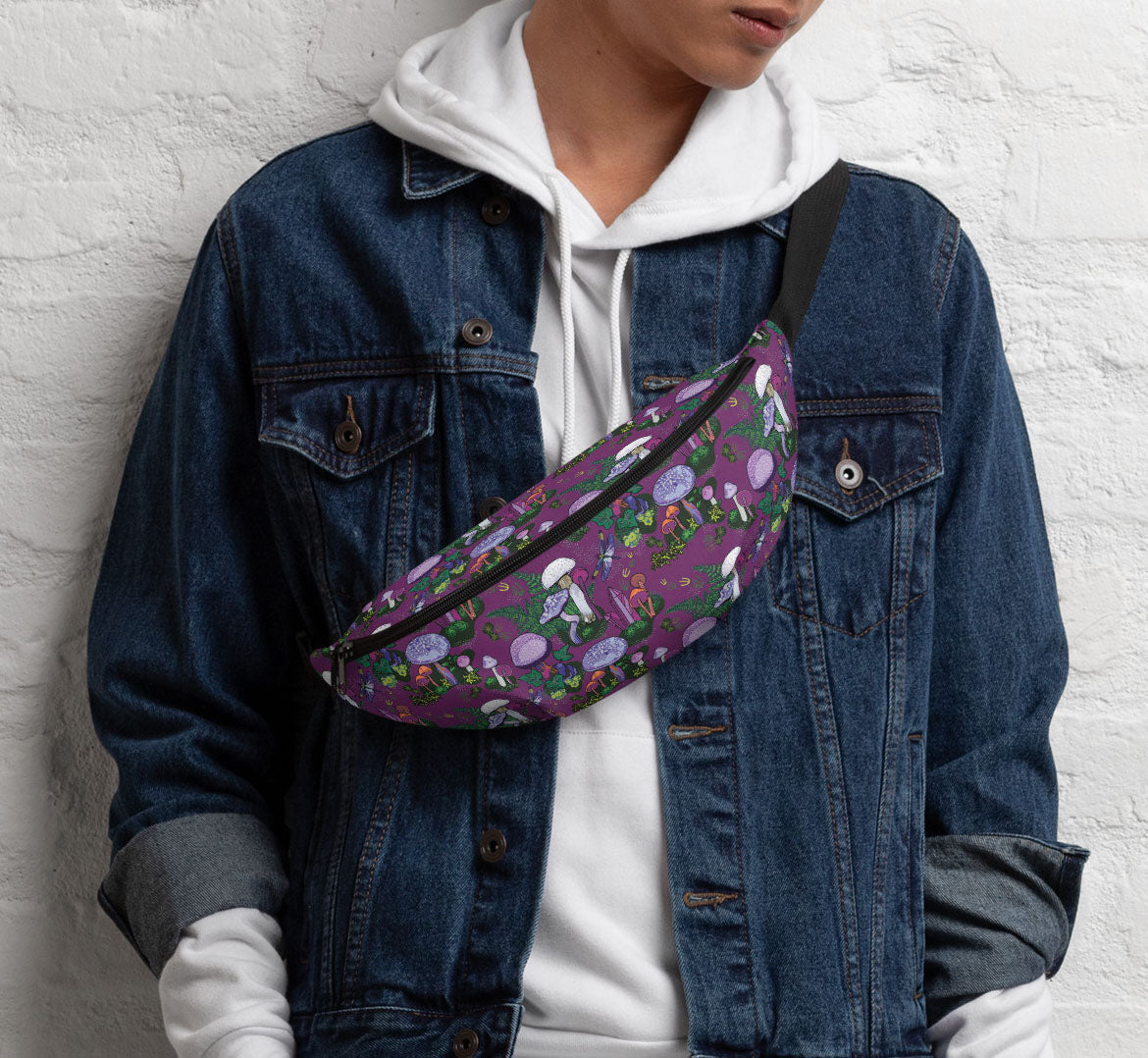 Woman standing against a white brick wall. She is wearing a white sweat shirt under a denim jacket. She is wearing the hip pack like a sling bag over her shoulder.