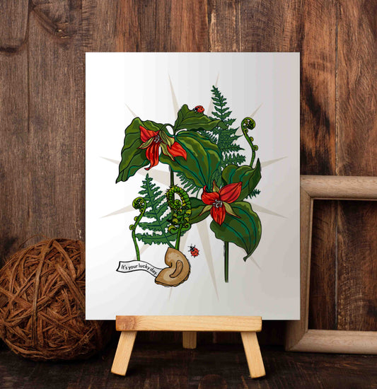 Trillium, Fern, and Fortune Cookie art poster on a wooden easel set against a wood-paneled wall. This hand-drawn illustration is designed to bless your space with good fortune. At the center, a fortune cookie declares that you are surrounded by blessings. Trillium flowers represent purity, balance, and the strength found in overcoming challenges. Ferns speak to growth, protection, and quiet magic. And the ladybugs? They bring a touch of luck and the promise of positive change.