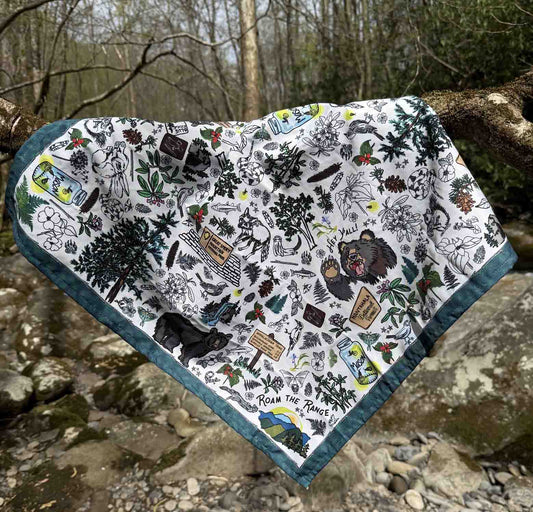 The Smoky Mountains bandana scarf is lying across a tree branch in the Smoky Mountains. You can see the detail of the illustrations on the bandana. The illustrations are iconic images which include a mason jar with fireflies, black bears, hiking symbols and trail markers, mountain laurel, wildflowers, foxes, waterfalls, pine trees and other plants.