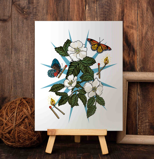 The Wild Rose and Butterflies art poster rests on a wooden easel, framed by a warm, wood-paneled wall. Set against a blue starburst, wild white roses symbolize love and spiritual growth. They're surrounded by butterflies and burning matches—symbols of transformation and the challenges we’ve overcome to be reborn.