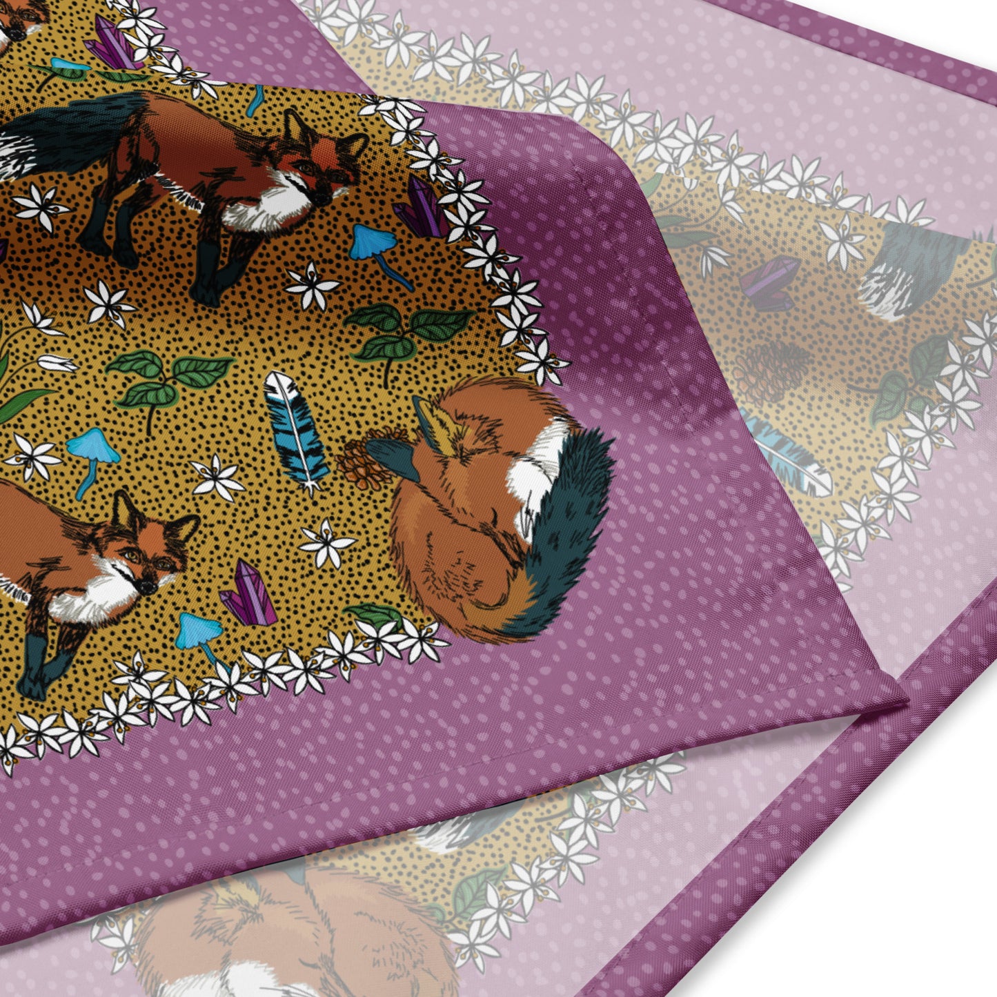 This is a close-up image of the bandana scarf folded over. It shows the pattern that includes foxes, crystals, feathers, pinecones, mushrooms, and flowers. The color palette is greens, turquoise blue, white, and a bit of brown.