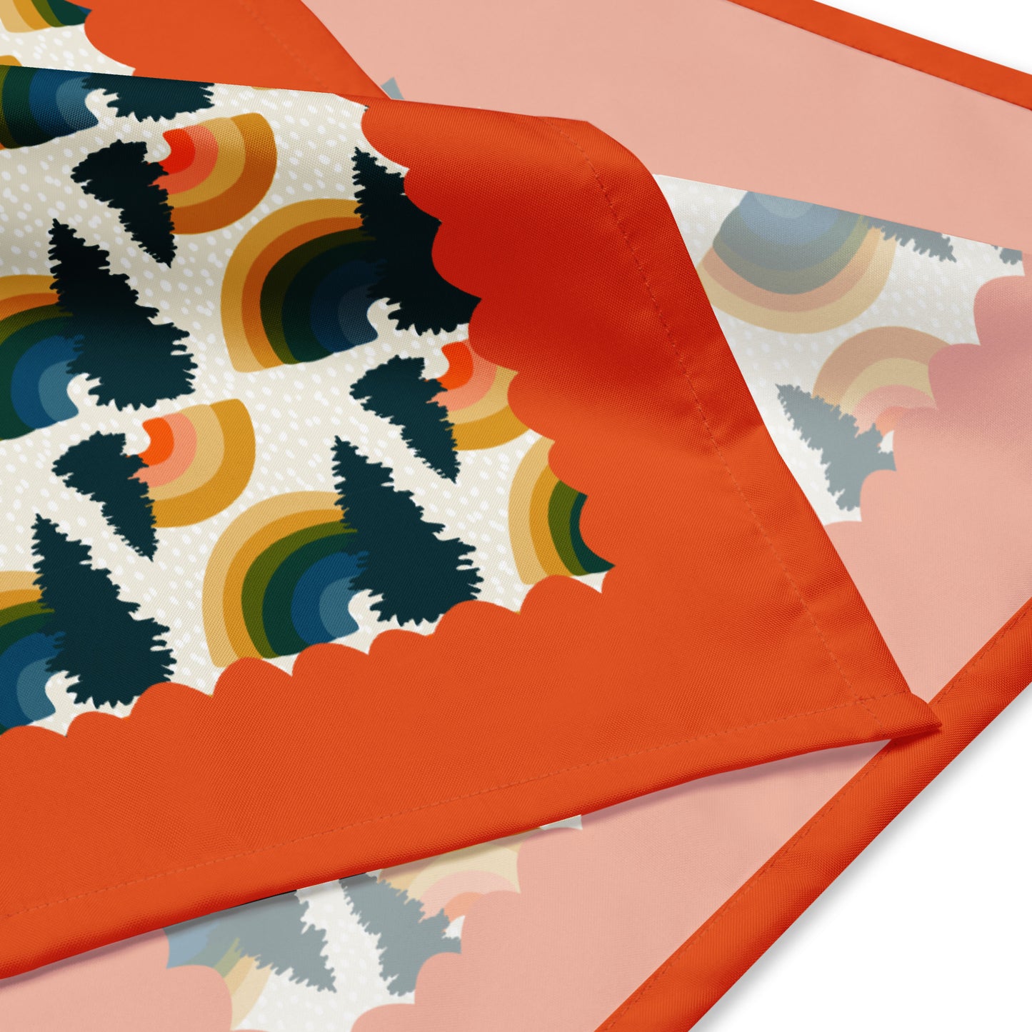 This is a close-up image of the bandana scarf folded over. It shows the pattern that includes mini rainbows and pine trees. There is a red scalloped border around the outside.