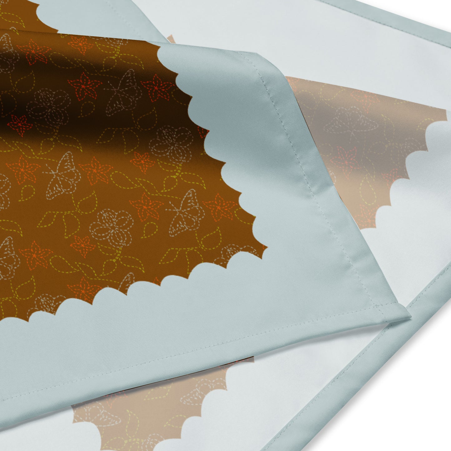 This is a close-up shot of the corner of the Cowgirl Stitches bandana. It shows a close-up of the pattern which is green leaves, butterflies, and flowers illustrated to look like stitches on a cowboy boot. The pattern has a warm brown background and is framed by a robin blue scalloped edge.