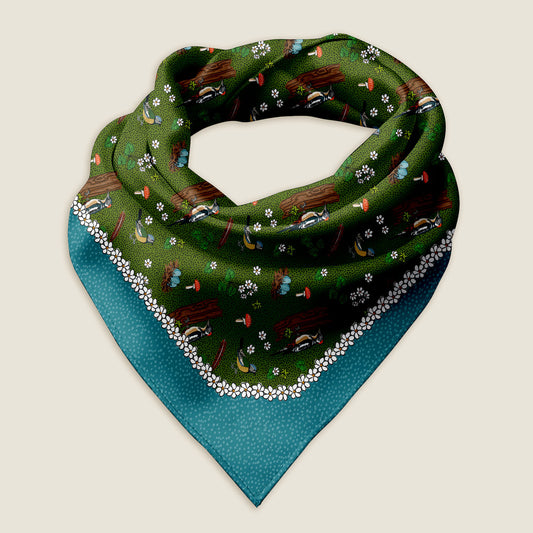 This image features the Bird & Mushrooms bandana scarf styled as a wrap against a soft ivory background. The design showcases a hand-drawn scene with a rich green center, where a woodpecker and other birds perch among nests, red mushrooms, and delicate white flowers. A white floral border frames the scene, followed by a deep blue edge accented with tiny white dots—an artful layering of nature and charm.