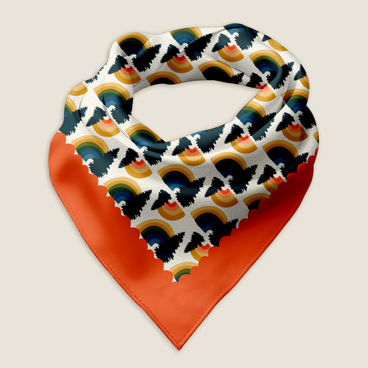 This is an image of the Rainbow Pine bandana scarf styled into a wrap showing the hand-drawn rainbows with pine tree on a white background with a scalloped red border. The words "Peace & Love" are tucked into one corner of the red border. Behind the scarf is a graphic of pine trees. The WanderWrap logo is at the top and the Posh Pad Designs logo is in the lower right corner. The Rainbow bandana is laying on an ivory background.