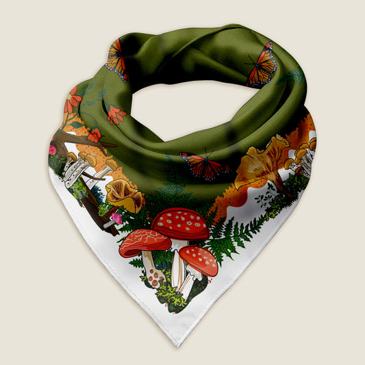 The red mushroom bandana on an ivory background shows the details of the hand-drawn artwork, which includes a variety of red, pink, and yellow mushrooms, butterflies, ferns, red flowers, and moss illustrated around a green middle with a white border.