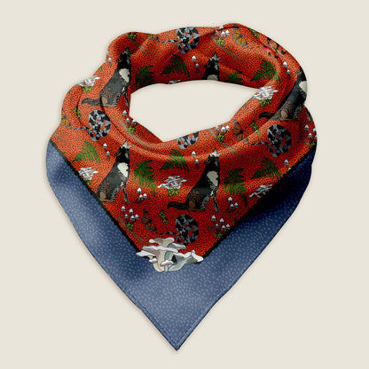 Wolf bandana scarf on an ivory background. This is an image of the Wild Wolf bandana scarf styled into a wrap showing the hand-drawn artwork of howling wolves, curly snakes, moss, mushrooms,  ferns, and butterflies on a red background with a warm purple border with a white dot pattern. 