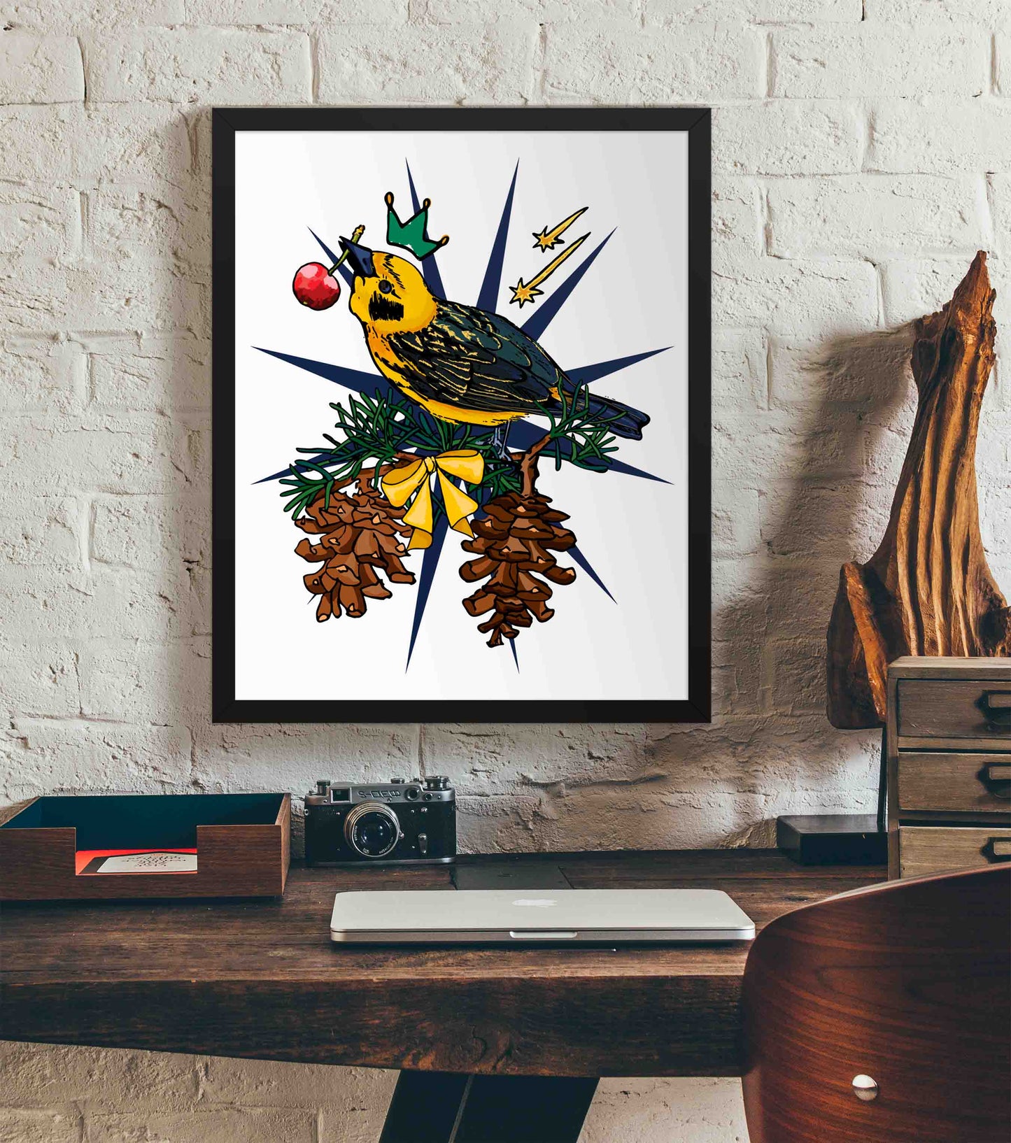 This photo shows the framed Crowned Golden Finch art print hanging on a white brick wall. Below it, there is a wood. The desk has a camera, laptop, and wood sculpture on it.