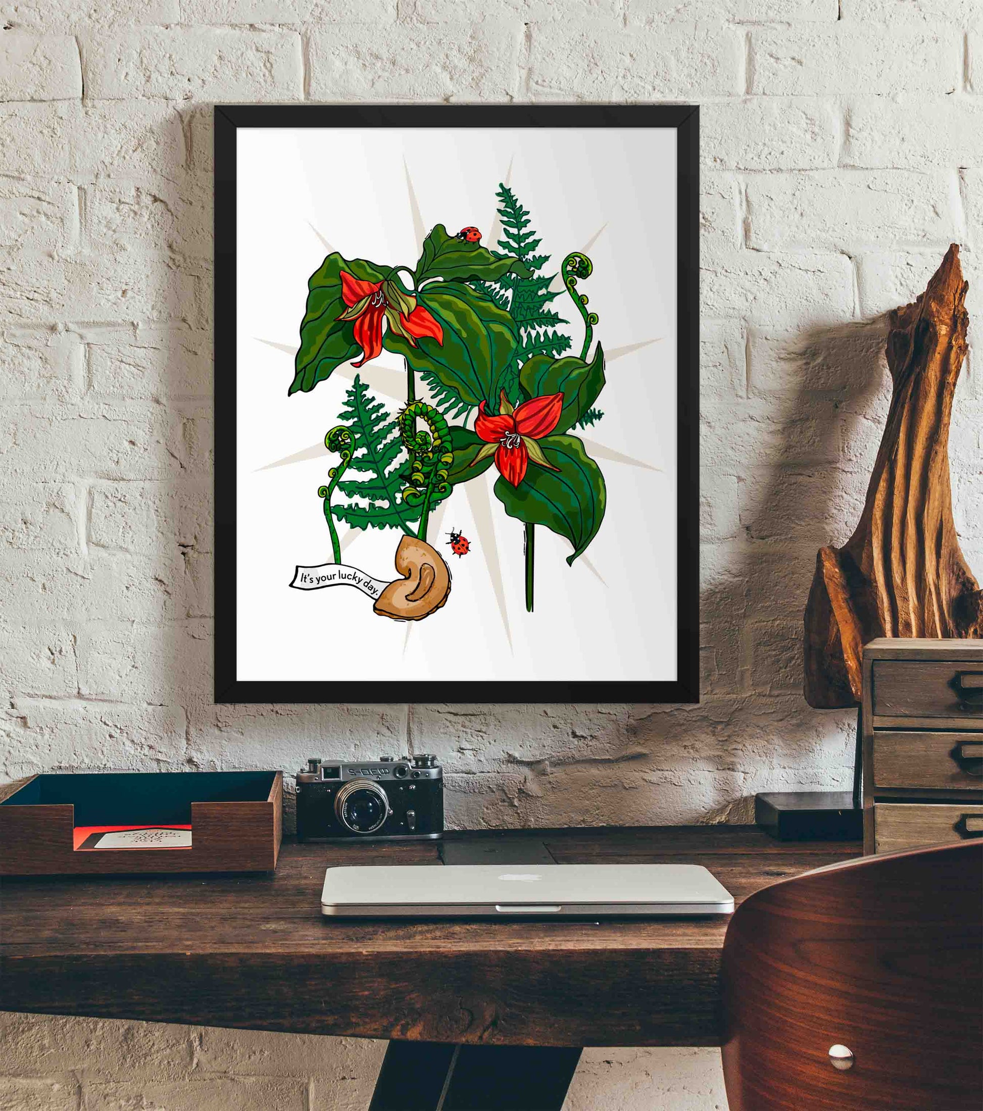 This photo shows the framed Red Trillium art print hanging on a white brick wall. Below it, there is a wood. The desk has a camera, laptop, and wood sculpture on it.