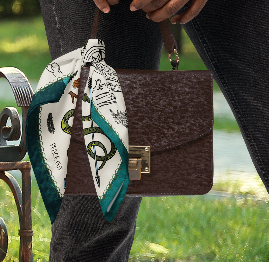 This is a photo of the bandana scarf tied to a small brown handbag that is being held by a woman in the park.