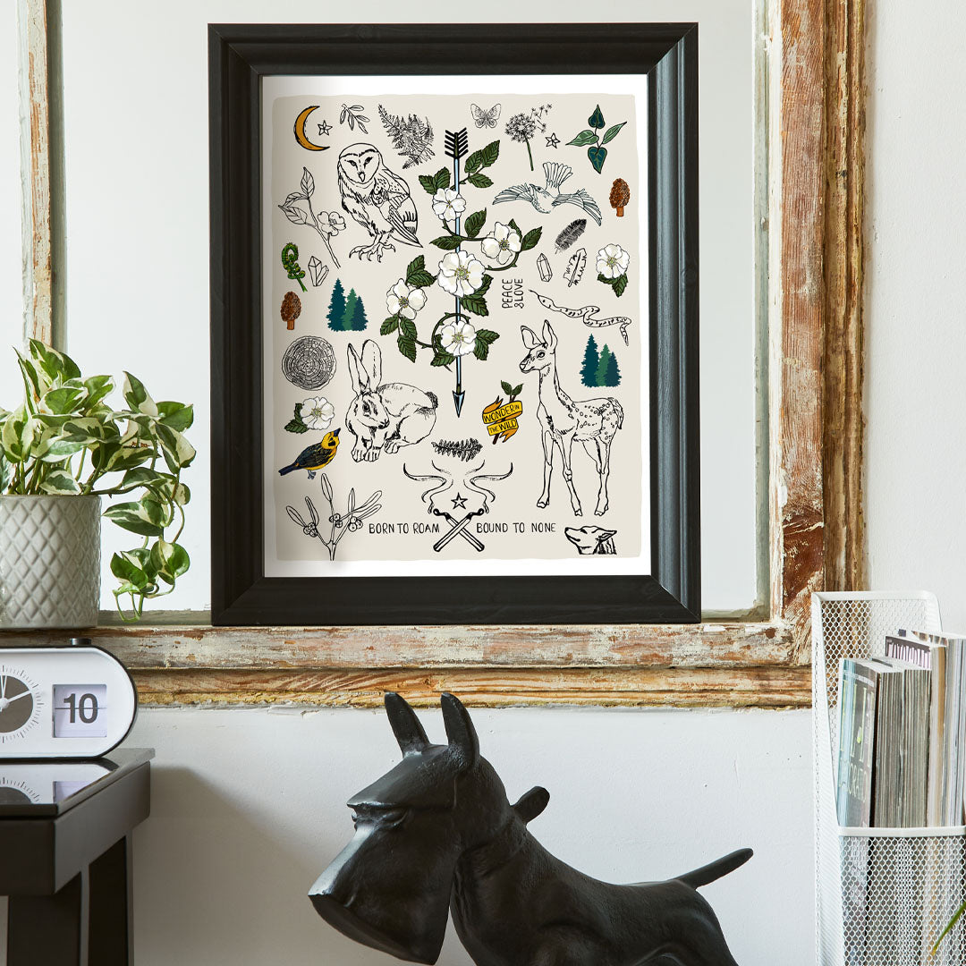 Born to Roam art print poster framed in a thick black frame in a home office. The artwork has drawings of wild roses, an owl, deer, rabbit, wolf, a crescent moon, ferns, crystals, mushrooms, dandelions, burning matches, and forest icons.