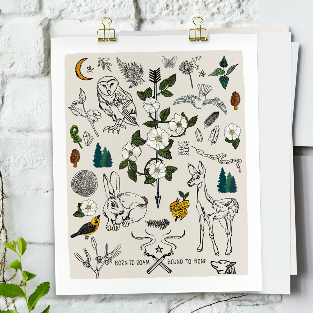 Born to Roam art print poster hung with gold clips on a white brick wall. The artwork has drawings of wild roses, an owl, deer, rabbit, wolf, a crescent moon, ferns, crystals, mushrooms, dandelions, burning matches, and forest icons.