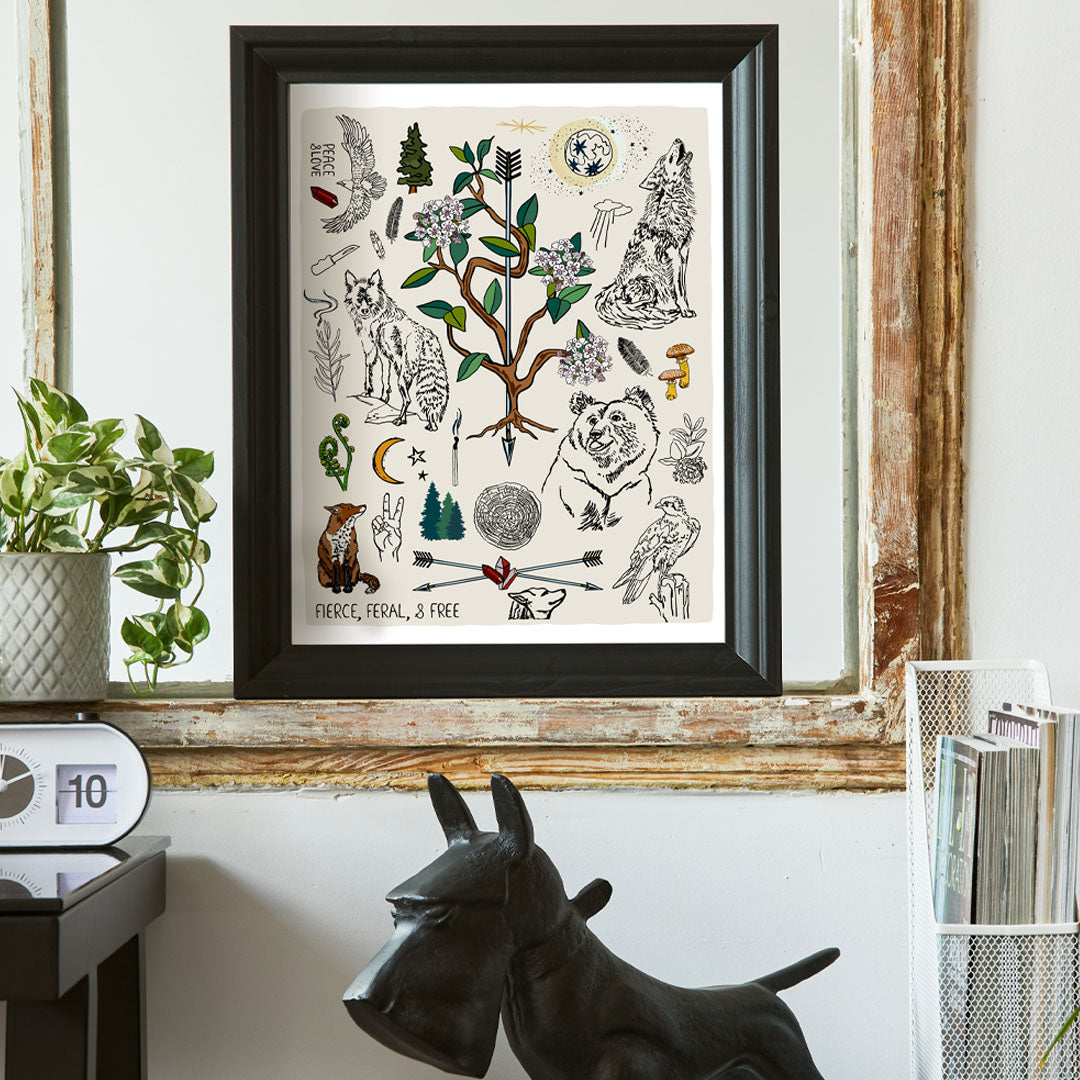 Fierce, Feral, and Free art print poster framed in a thick black frame in a home office. The artwork has bears, wolves, birds, forest icons, a full moon, a crescent moon, crossed arrows, mushrooms, burning sage, matches, and a camp knife. 
