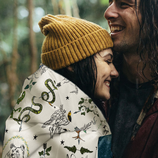 Two people embracing outdoors. The young woman is wearing a mustard yellow beanie, with the Fierce, Feral, and Free sherpa blanket draped around her shoulders.