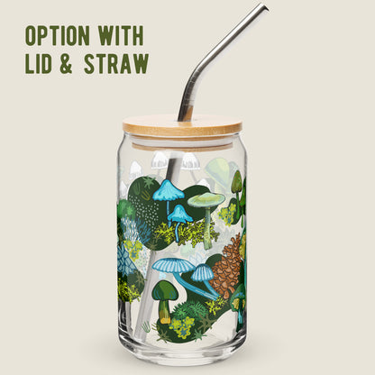 The Green Mushroom glass tumbler with the bamboo lid and metal straw option on an ivory background. The artwork on the glass is illustrations of various green and blue mushrooms, green leaves, moss, alongside pinecones and moths. The color palette is a mix of vibrant mossy greens and  earthy blues.
