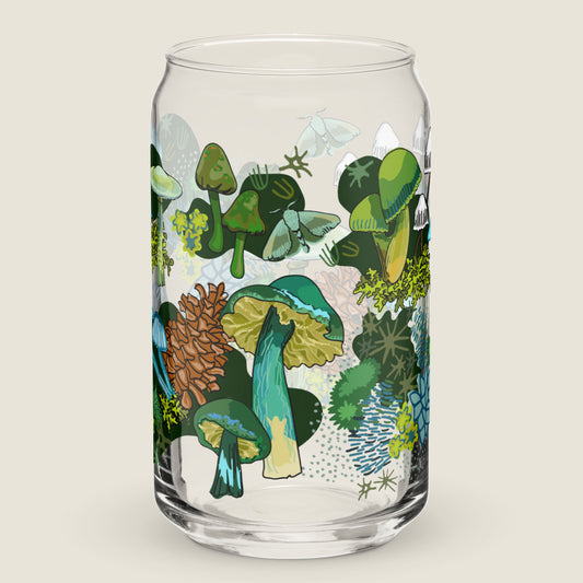 The Green Mushroom glass tumbler on an ivory background. The artwork on the glass is illustrations of various green and blue mushrooms, green leaves, moss, alongside pinecones and moths. The color palette is a mix of vibrant mossy greens and  earthy blues.