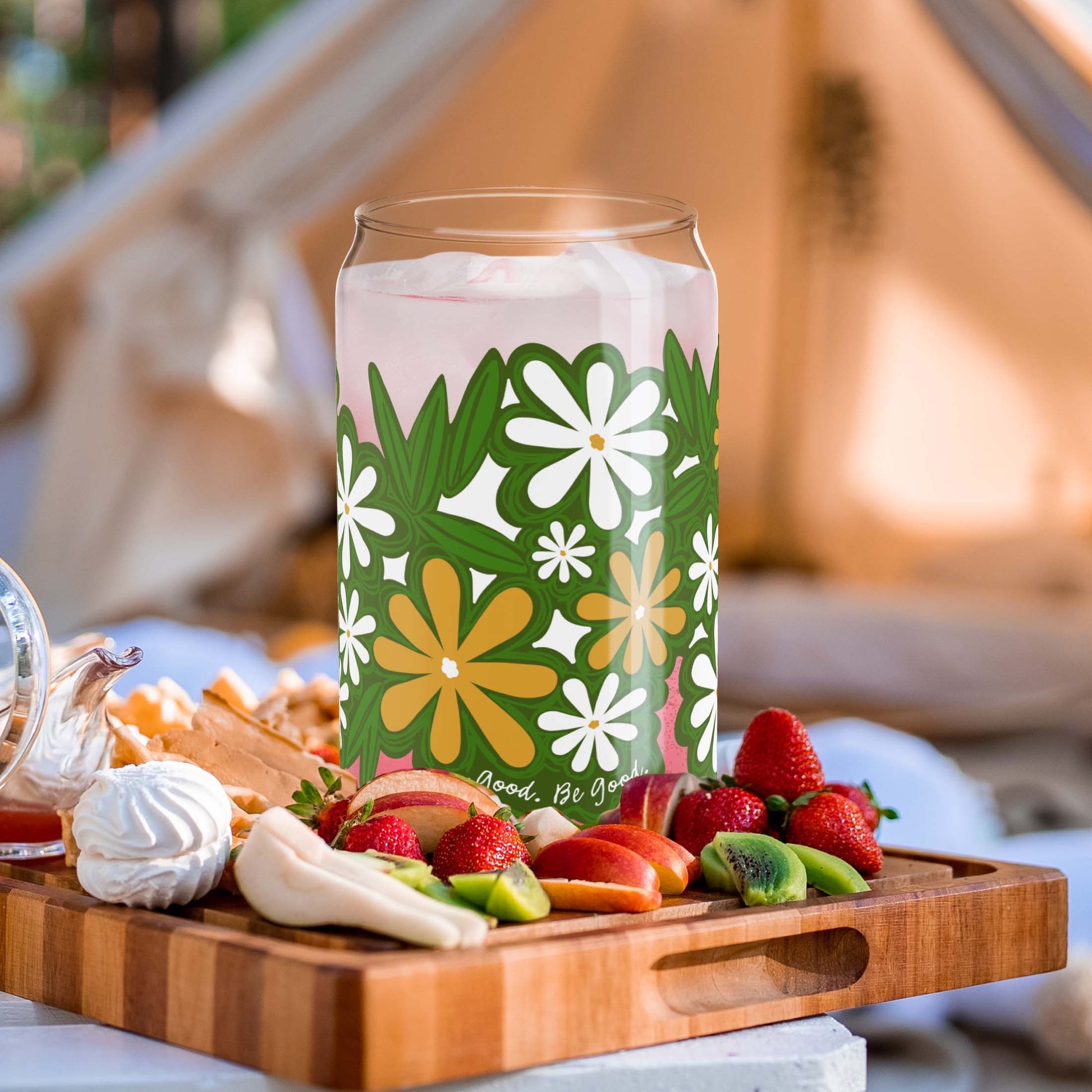 This is a boho-style picnic scene with the Daisy Flower glass filled with a pink drink and ice. It's sitting on a wooden cutting board with fruit. In the background there is a bohemian-style tent and trees.
