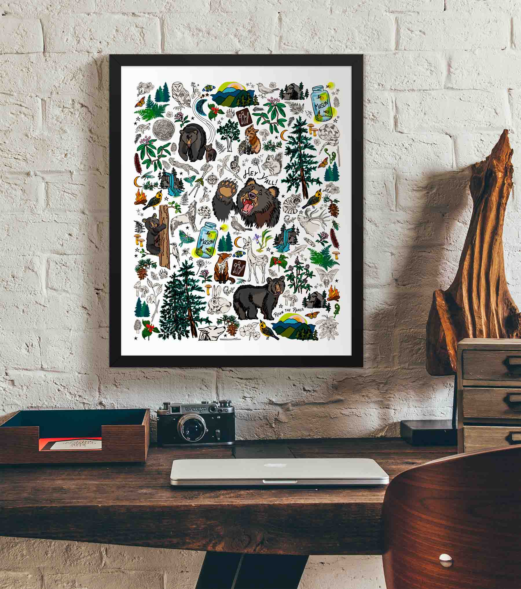 This photo shows the framed Mountain Hikes art print hanging on a white brick wall. Below it, there is a wood. The desk has a camera, laptop, and wood sculpture on it.