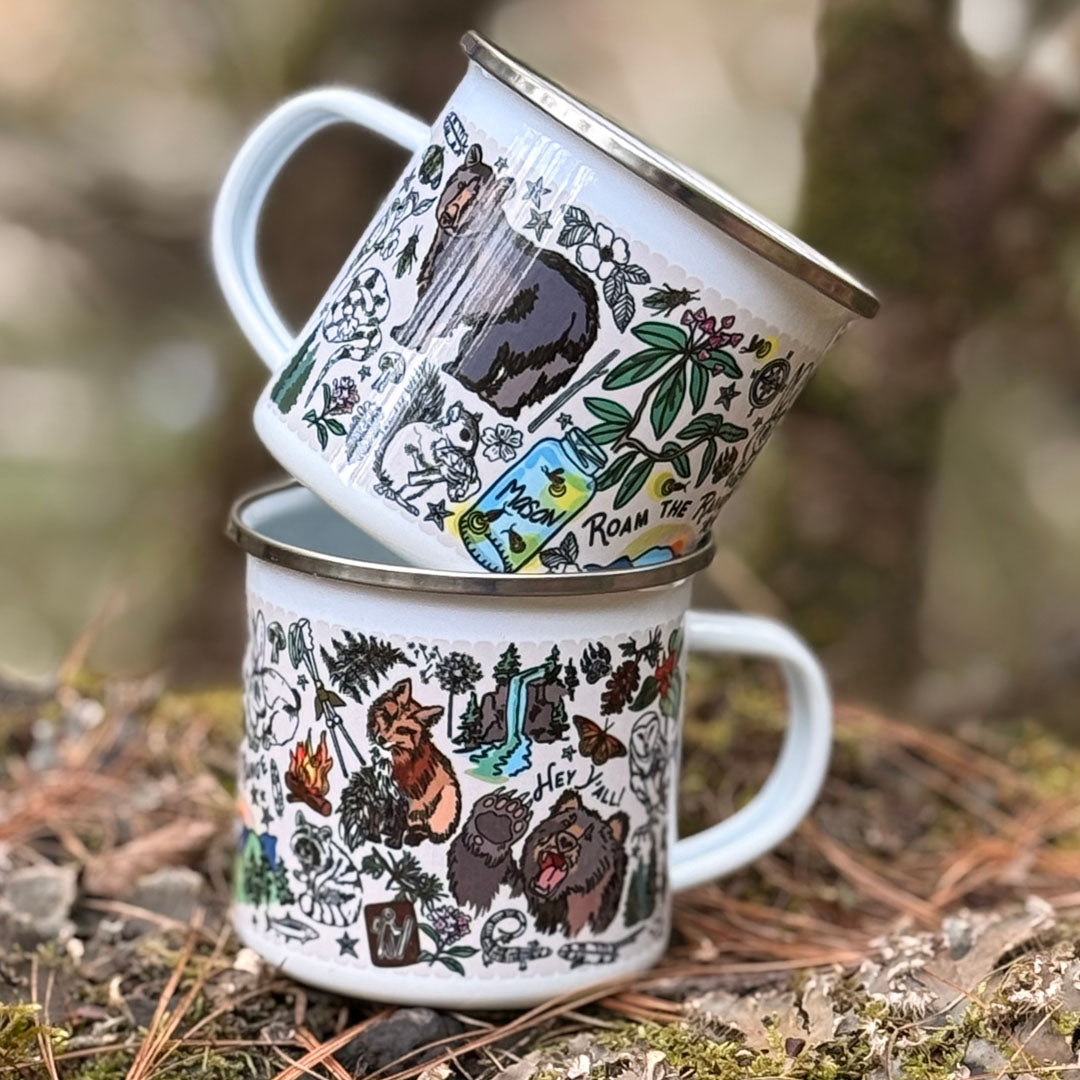 Two Mountain Hikes enamel camp mugs with nature-themed designs stacked on a forest floor.