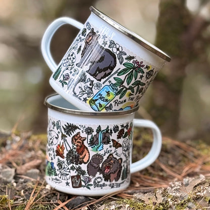 Two Mountain Hikes enamel camp mugs with nature-themed designs stacked on a forest floor.