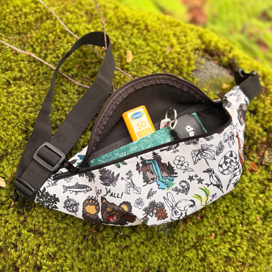 Mountain Hikes hip pack laying on a moss log. The zipper is open to show the inside where there are keys and sunscreen poking out.