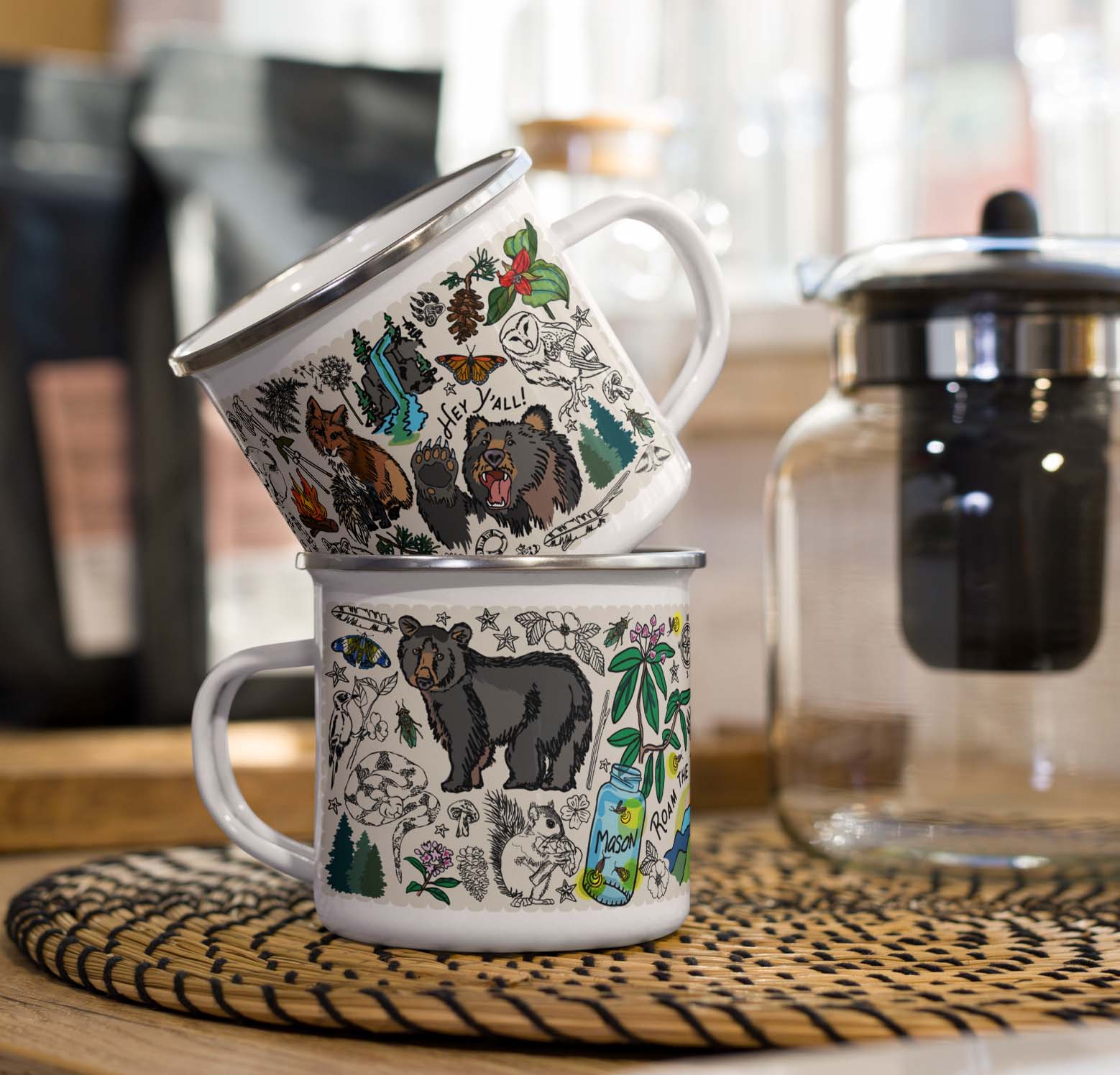 Two Mountain Hikes metal enamel mugs with nature-themed designs stacked on a kitchen counter beside a coffee pot.