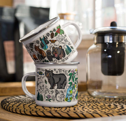 Two Mountain Hikes metal enamel mugs with nature-themed designs stacked on a kitchen counter beside a coffee pot.