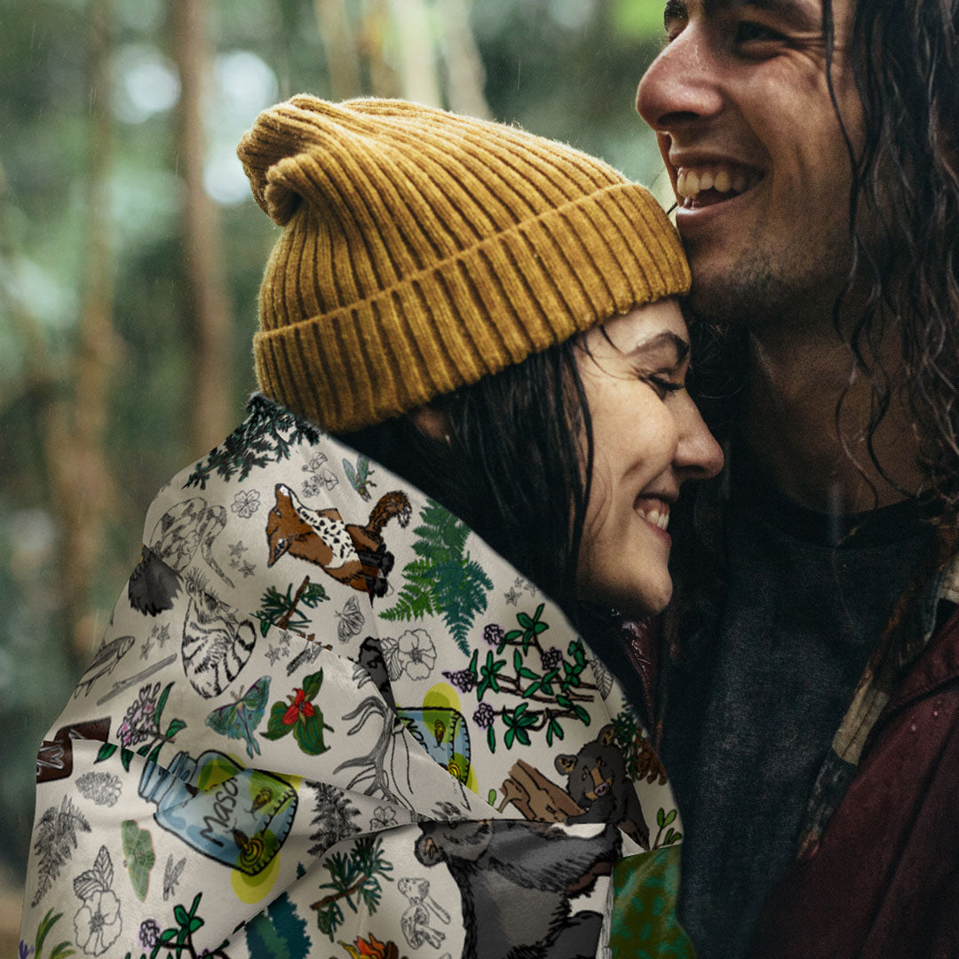 Two people embracing outdoors. The young woman is wearing a mustard yellow beanie, with the Mountain Adventures sherpa blanket draped around her shoulders.