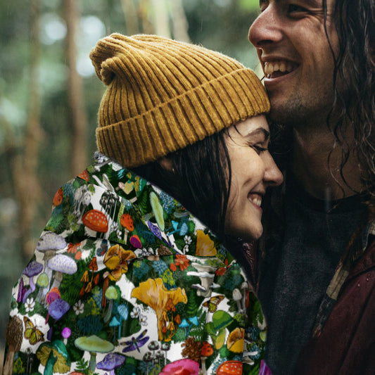 Two people embracing outdoors. The young woman is wearing a mustard yellow beanie, with the Wild Mushrooms sherpa blanket draped around her shoulders.