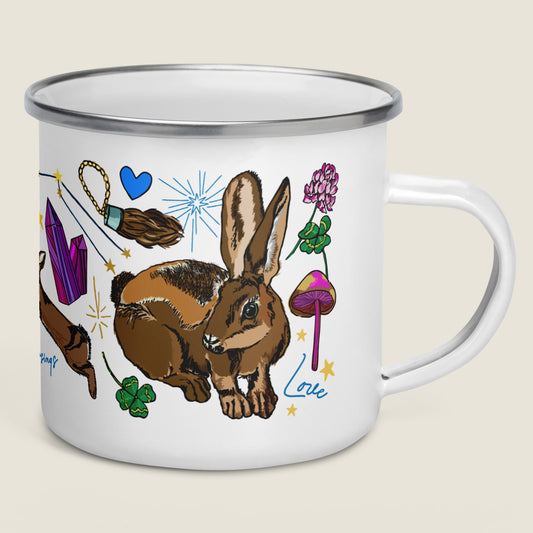 Orion Constellation enamel camp mug on an ivory background. The artwork has illustrations of good luck charms, like lucky rabbit's foot, four-leaf clovers, mystical mushrooms, blue hearts, stars, and crystals.