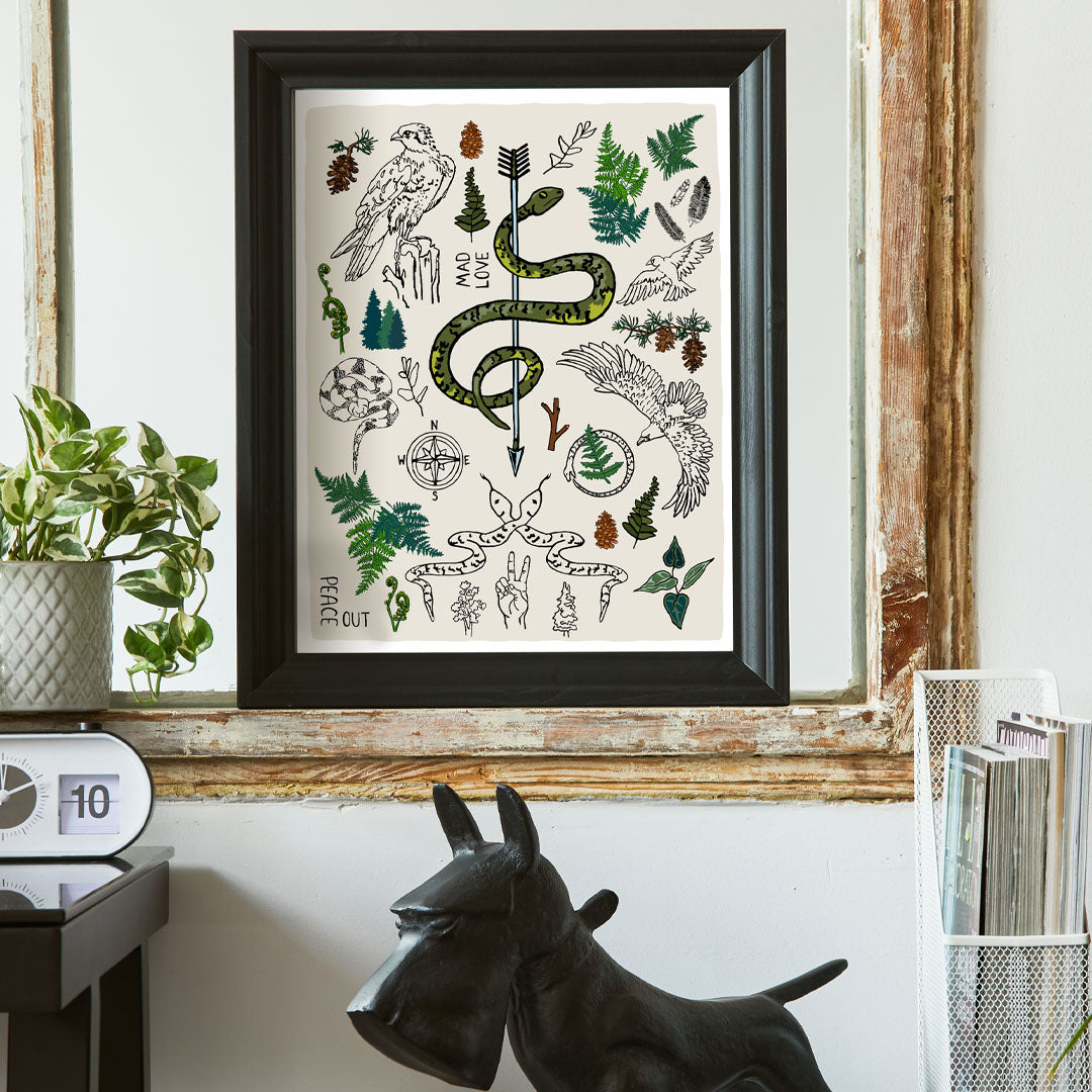 Fierce, Feral, and Free art print poster framed in a thick black frame in a home office. The artwork has illustrations of eagles, snakes, ferns, a north star compass, pinecones, pine trees, an arrow, feathers, peace sign, and forest icons. 