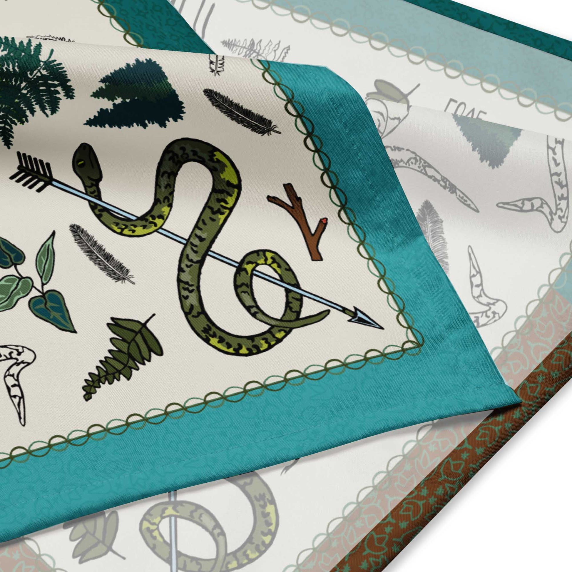 Close up detail of the bandana scarf. It shows the illustrated pattern of snakes entwined with an arrow, feathers, leaves, pinecones, and forest motifs.